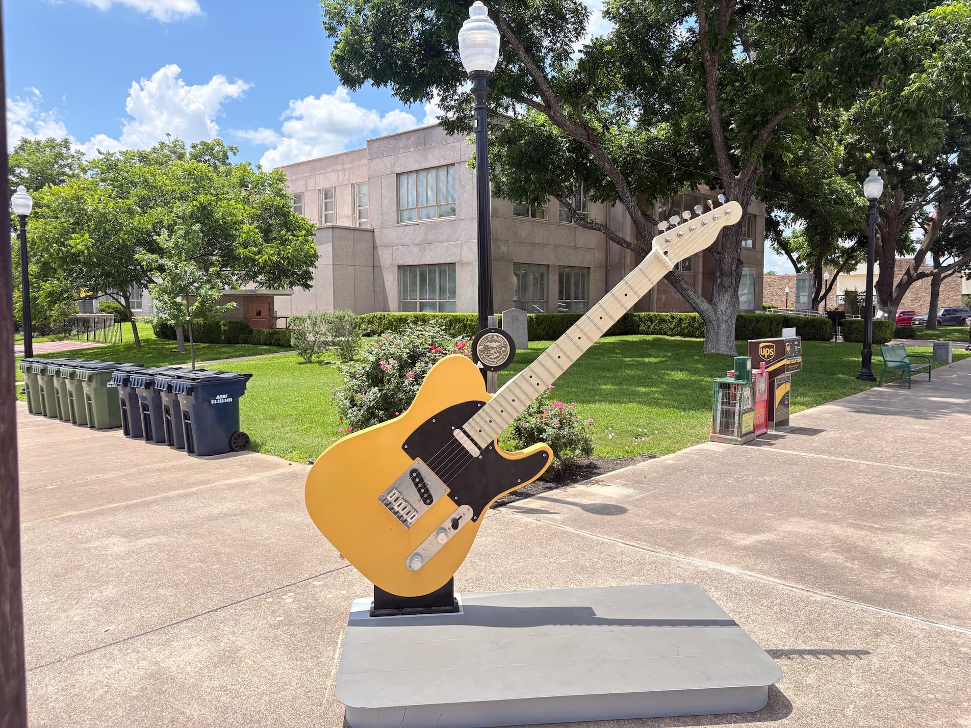 Large yellow electric guitar sculpture on a raised platform in a sunny campus-like courtyard with trees, benches, and a beige building in the background